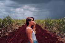 Бічний вид на жінку в повсякденному одязі стоїть біля коричневого ґрунту серед зелених тропічних рослин під сірим хмарним небом на Кубі. — стокове фото