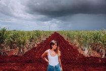 Женщина-путешественница в повседневной одежде стоит на перекрестке с коричневой почвой среди зеленых тропических растений под серым облачным небом на Кубе — стоковое фото