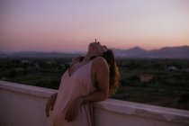 Side view of peaceful woman lying with closed eyes on fence of balcony and smoking cigarette with sunset on blurred background — Stock Photo