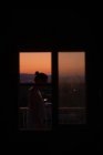 Вид збоку на молоду безлику леді, що стоїть на балконі з чудовим заходом сонця на розмитому фоні — стокове фото