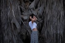 Vue latérale de la jeune brune en t-shirt blanc et jupe légère debout et fixant les cheveux avec des plantes tropicales sèches sur le fond — Photo de stock