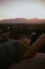 Vista lateral de la mujer tranquila acostada con los ojos cerrados en la cerca del balcón y fumar cigarrillo con puesta de sol sobre fondo borroso - foto de stock