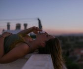 Вид збоку мирної жінки, що лежить із закритими очима на паркані балкона та курить сигарету із заходом сонця на розмитому фоні — стокове фото