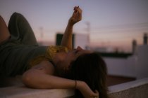 Vista lateral da mulher pacífica deitada com os olhos fechados na cerca da varanda e fumar cigarro com o pôr do sol no fundo turvo — Fotografia de Stock