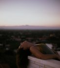Peaceful woman leaning with closed eyes on fence of balcony with sunset on blurred background — Stock Photo
