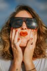 Stilvolle braune gelockte Frau mit rotem Lippenstift in trendiger Sonnenbrille blickt in die Kamera, während sie das Gesicht in die Handflächen presst — Stockfoto