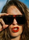 Elegante mujer de pelo rizado marrón con lápiz labial rojo en gafas de sol de moda mirando a la cámara mientras hace caras - foto de stock