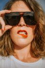 Стильная брюнетка с кудрявыми волосами и красной помадой в модных солнцезащитных очках, смотрящая в камеру, делая лица — стоковое фото