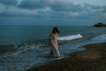 Voyageuse pieds nus en robe légère marchant parmi les petites vagues de la mer sur le littoral vide au crépuscule en regardant loin — Photo de stock