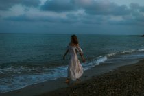 Босонога жінка мандрівник у легкій сукні, що йде серед малих морських хвиль на порожній береговій лінії в сутінках, дивлячись далеко — стокове фото
