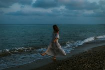 Viajante mujer descalza vestida de vestido ligero caminando entre pequeñas olas marinas en la costa vacía al atardecer mirando hacia otro lado - foto de stock