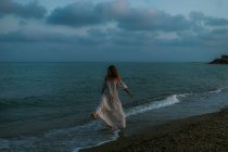 Босонога жінка мандрівник у легкій сукні, що йде серед малих морських хвиль на порожній береговій лінії в сутінках, дивлячись далеко — стокове фото