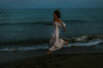 Босонога жінка-мандрівник у легкій сукні, що бігає серед невеликих морських хвиль на порожній береговій лінії в сутінках, дивлячись далеко — стокове фото