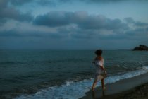 Просмотр анонимных босых женщин-путешественниц в светлых платьях, танцующих среди маленьких морских волн на пустой береговой линии в сумерках, смотрящих в сторону — стоковое фото