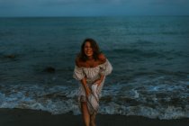 Босоногая женщина-путешественница в легком платье танцует среди маленьких морских волн на пустой береговой линии в сумерках, глядя в камеру — стоковое фото