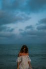 Женщина в светлом платье гуляет среди маленьких морских волн на пустой береговой линии в сумерках, глядя вниз — стоковое фото