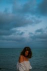 Mulher desfocada em vestido claro andando entre pequenas ondas do mar na costa vazia ao anoitecer olhando para baixo — Fotografia de Stock
