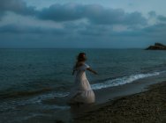 Back view anonymous of barefoot woman traveler in light dress dancing among small sea waves on empty coastline at dusk looking away — Stock Photo