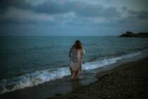 Barfüßige Reisende in hellem Kleid spazieren zwischen kleinen Meereswellen an der leeren Küste in der Abenddämmerung und schauen weg — Stockfoto