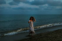 Босонога жінка мандрівник у легкій сукні танцює серед малих морських хвиль на порожній береговій лінії в сутінках, дивлячись на камеру — стокове фото