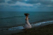 Вид збоку анонімний босоніж жінка мандрівник у легкій сукні танцює серед невеликих морських хвиль на порожній береговій лінії в сутінках, дивлячись далеко — стокове фото