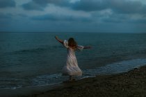 Voltar vista anônimo de mulher descalça viajante em vestido leve dançando entre pequenas ondas do mar na costa vazia ao entardecer olhando para longe — Fotografia de Stock