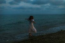 Вид ззаду анонімний босоніж жінка мандрівник у легкій сукні танцює серед невеликих морських хвиль на порожній береговій лінії в сутінках, дивлячись далеко — стокове фото