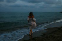 Back view of anonymous barefoot woman traveler in light dress running among small sea waves on empty coastline at dusk — Stock Photo