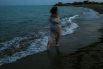 Visão traseira de mulher anônima descalça viajante em vestido leve correndo entre pequenas ondas do mar na costa vazia ao entardecer — Fotografia de Stock