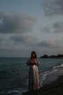 Жінка-мандрівник у легкій сукні, що йде серед невеликих морських хвиль на порожній береговій лінії в сутінках, дивлячись на камеру — стокове фото