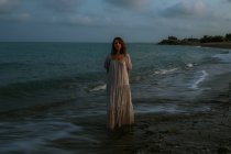 Viajante mujer descalza vestida de vestido ligero caminando entre pequeñas olas marinas en la costa vacía al atardecer mirando la cámara - foto de stock
