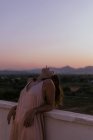 Side view of peaceful woman lying with closed eyes on fence of balcony and smoking cigarette with sunset on blurred background — Stock Photo