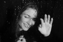 Blanco y negro de mujer sonriente de pie detrás del vidrio en gotas de agua tocando la superficie con los ojos cerrados - foto de stock