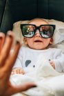 From above of newborn infant in pajama with open mouth in sun glasses lying on bed at home — Stock Photo