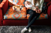 Сумна новонароджена дитина в білій піжамі на руках доглядаючої матері з схрещеними ногами сидить на ліжку біля собак вдома — стокове фото