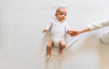 Вид зверху на безлику жінку, яка торкається рук радісного новонародженого немовляти з відкритим ротом у білій піжамі, розважається лежачи на ліжку, дивлячись геть — стокове фото