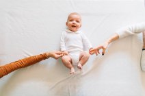 Vista dall'alto di donne senza volto che toccano le mani di gioioso neonato con la bocca aperta in pigiama bianco divertirsi sdraiato sul letto guardando la fotocamera — Foto stock