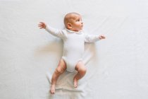 Vista superior de un precioso bebé en pijama blanco con la boca abierta acostado en la cama moviendo los brazos y las piernas mirando hacia arriba - foto de stock