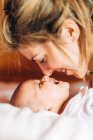 Closeup of glad mom touching nose by nose of baby playing with newborn infant with open mouth having fun lying on bed at home — Stock Photo
