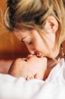 Крупный план радостной мамы, касающейся носа за носом ребенка, играющей с новорожденным младенцем с открытым ртом, веселящимся лежа на кровати дома — стоковое фото