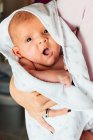 Крупный план спокойного новорожденного младенца в белом одеяле на руках урожая заботливой матери дома, смотрящей в камеру — стоковое фото