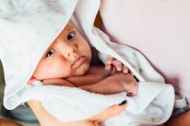 Крупним планом спокійна новонароджена дитина в білій ковдрі на руках доглядаючої матері вдома дивлячись на камеру — стокове фото
