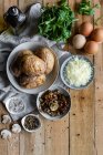 Von oben Schüsseln mit Brauen gefüllte Kartoffeln grüne Kräuter gebratene Champignons Eier geriebenen Käse und Flasche mit Olivenöl auf Holztisch — Stockfoto