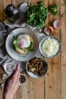 Top view of crop hand with knife cutting fried egg on potato on wooden table with fried mushrooms grated cheese and herbs — Stock Photo
