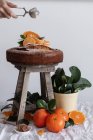 Cultivez une personne anonyme en versant de la poudre de sucre avec une passoire à thé ronde en métal au-dessus d'un gâteau appétissant frais sur des tabourets en bois entourés de mandarine mûre orange et de plantes vertes en pot — Photo de stock