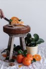Cultive pessoa anônima derramando açúcar em pó com metal coador de chá redondo acima do bolo apetitoso fresco em banquetas de madeira cercadas por tangerina madura laranja e planta verde em panela — Fotografia de Stock