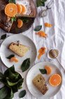 Сверху аппетитный сладкий торт и спелый апельсиновый мандарин режут и подают на белых тарелках на столе, украшенном растениями — стоковое фото