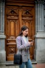 Charming young lady in stylish casual outfit smiling and looking away while surfing on mobile phone in city street — Stock Photo