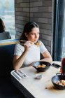 Von oben eine nachdenkliche schwarzhaarige Frau in lässiger Kleidung, die Essstäbchen benutzt und asiatisches Essen mit Gemüse isst, während sie durch ein Fenster im Café blickt — Stockfoto