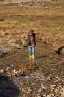 Adorable enfant en bottes en caoutchouc jaune debout dans une petite rivière au pied de montagnes enneigées pierreuses par temps lumineux — Photo de stock